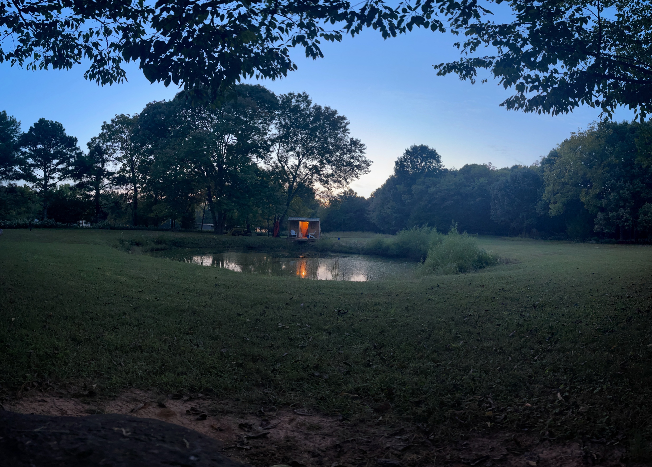 Atmospheric dusk scene of large mowed property with pond and lit dock reflecting in water